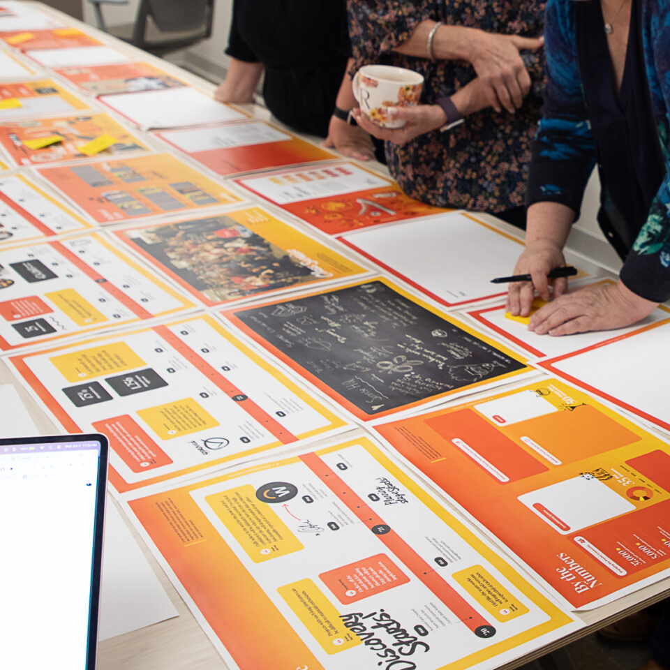 Three people stand at a table covered with colorful printed materials and posters. One person holds a mug, and another writes notes. A laptop is visible in the corner of the image.
