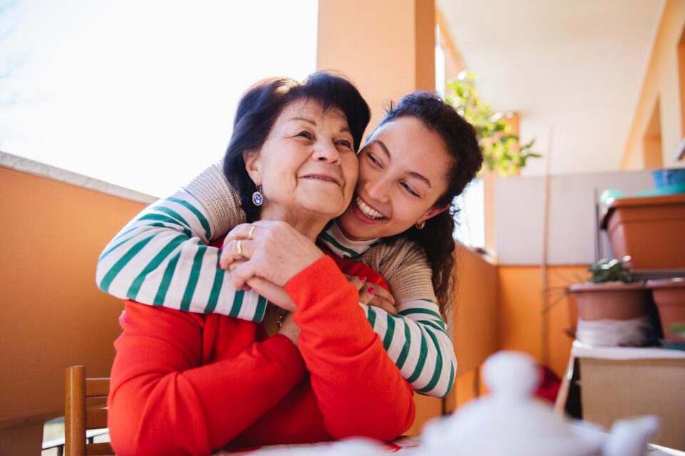 A young woman hugs an older woman from behind, both smiling happily while sitting outside on a sunny balcony with potted plants in the background.