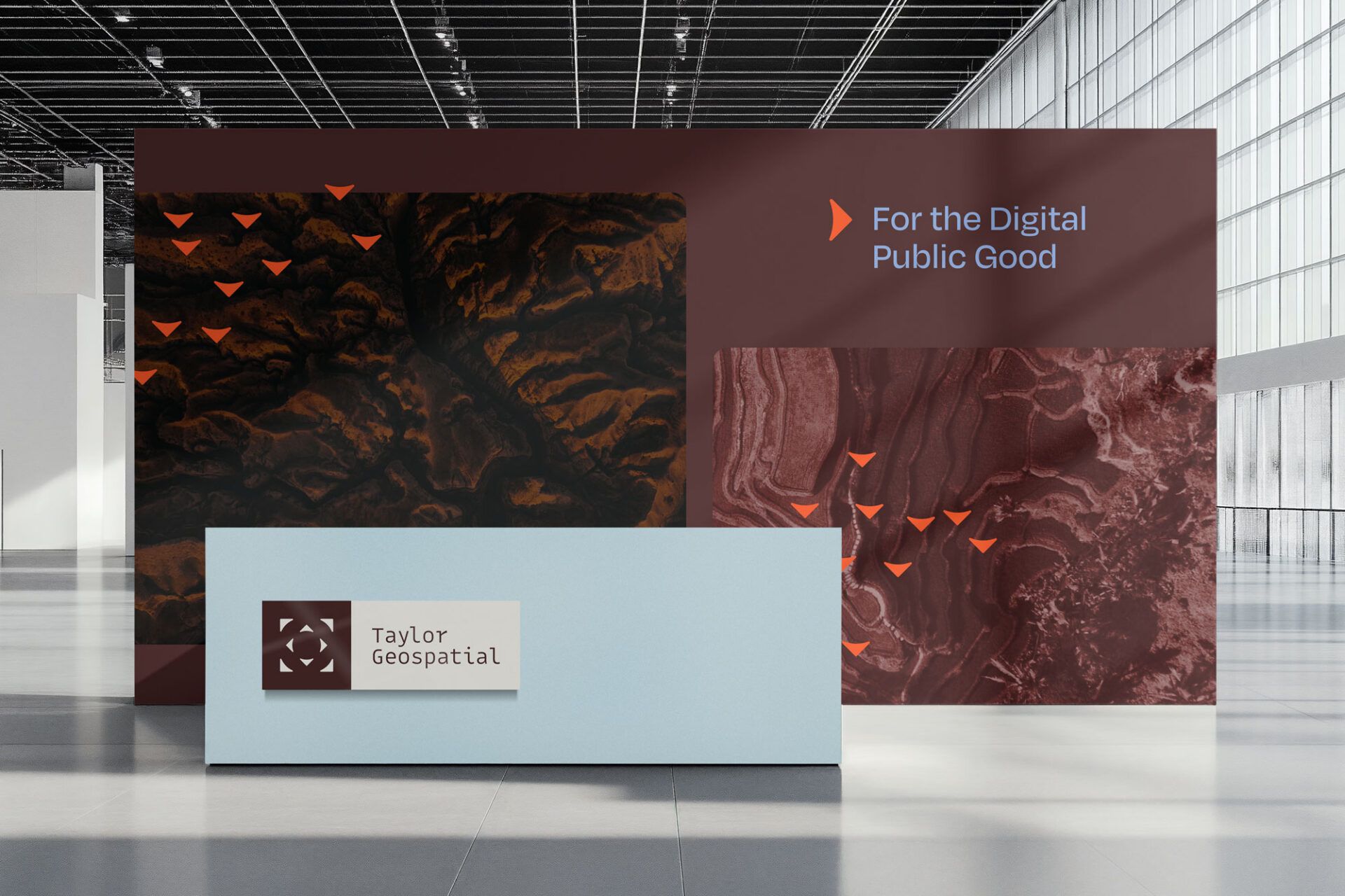 taylor-geospatial-reception-office-signage - Sprehe Studio Exhibition wall with layered burgundy and blue panels, abstract textures, orange triangles, and the text “For the Digital Public Good.” A light blue panel toward the front bears the Taylor Geospatial logo.