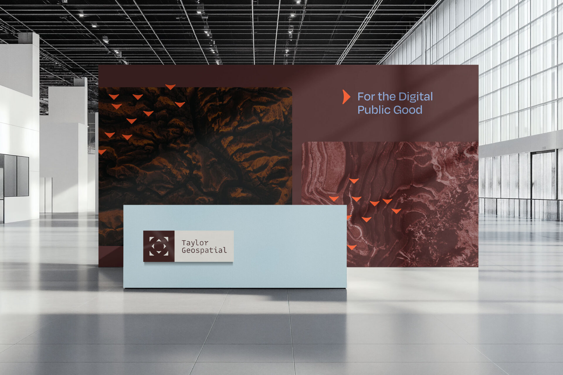 A modern exhibition booth with a light blue desk labeled Taylor Geospatial and a large backdrop displaying abstract brown patterns and the text For the Digital Public Good in a spacious, sunlit hall.