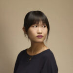 A woman with straight black hair and bangs, wearing a dark top and a gold necklace, looks to her left against a plain beige background.