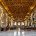 A grand library hall with ornate, gold-patterned ceiling, large arched windows, elegant chandeliers, wooden tables and chairs, and marble flooring. Natural light fills the spacious, historic room.