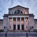 Exterior view of a grand neoclassical building with tall columns, statues, and detailed carvings above the entrance, photographed at dusk with lights illuminating the facade.