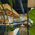 A modern restaurant interior with a colorful tropical mural, leafy green plants, a well-lit bar with shelves of bottles, blue accents, and cozy booth seating along the wall.